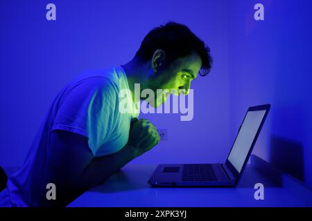 Ein passionierter Gamer sitzt am Schreibtisch und lehnt sich vor Spannung nach vorne, während er auf ein Notebook starrt. Der Raum ist mit einem fesselnden blauen Leuchten beleuchtet, en Stockfoto