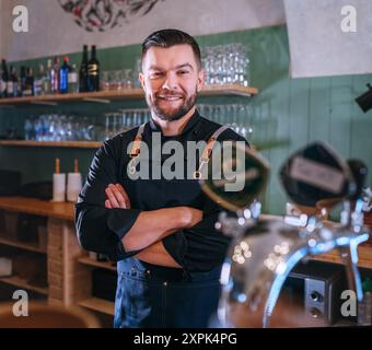 Porträt eines fröhlich lächelnden bärtigen Barkepers in schwarzer Uniform mit Schürze am Bartheken mit gezapftem Bier. Erfolgreiche Mitarbeiter, harte Arbeit, Stockfoto
