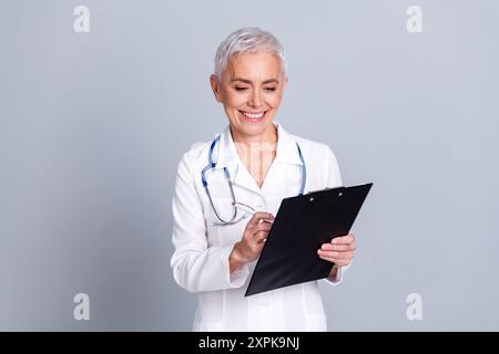 Foto der attraktiven älteren Frau Krankenhaus Arzt Arbeiter Schreiben Diagnose gekleidete weiße Gewand Uniform isoliert auf grauem farbigen Hintergrund Stockfoto