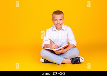 Foto des süßen intelligenten Jungen tragen weißes Hemd sitzen Boden schreiben Heimatmaske isoliert auf gelbem Hintergrund Stockfoto