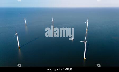 Sanfte Wellen schlagen gegen hoch aufragende Windmühlen und reflektieren in ruhigen Gewässern, während ein kleines Segelboot friedlich unter dem dunklen Himmel gleitet. Konzept der grünen Energie Stockfoto