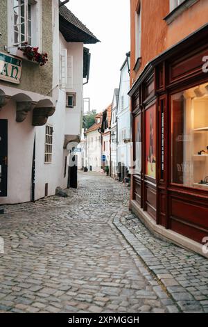 Eine malerische Kopfsteinpflasterstraße schlängelt sich durch eine historische europäische Stadt, gesäumt von malerischen Gebäuden in verschiedenen Farben. Das warme Licht von Shop Windo Stockfoto