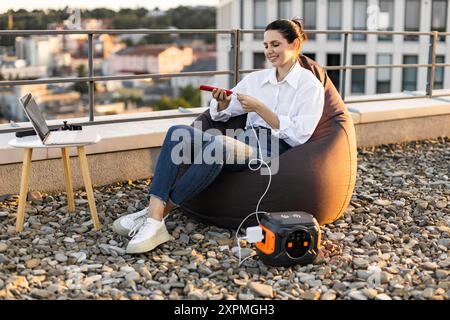 Frau, die Telefon mit tragbarem Kraftwerk auf dem Dach auflädt Stockfoto