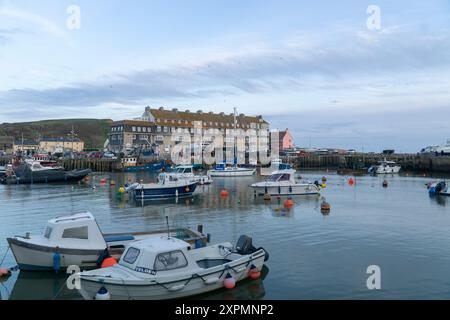 Kleine Boote, die im ruhigen Hafen von West Bay in Dorset verankert sind, mit Gebäuden am Wasser, Reflexionen und sanftem Tageslicht, die eine friedliche Szene schaffen Stockfoto