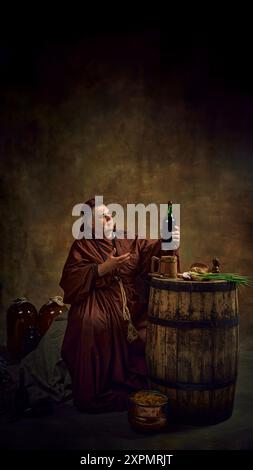 Mittelalterlicher Mönch, Mann, der von großen Gläsern Wein und Holzfässern Bier umgeben sitzt, eine Flasche Bio-Rotwein herstellt. Weinbautradition Stockfoto