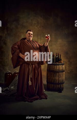 Stolzer Mann, mittelalterlicher Mönch in historischen Gewändern, der ein Glas Bier oder Cognac vor Vintage-Hintergrund mit Holzfass und Flasche oben anhebt. Stockfoto