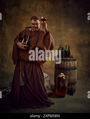 Mittelalterlicher Mönch in alter Kleidung hält Glas mit Cognac, Bier, Ale wieder Vintage Keller mit Holzfass und Flaschen oben. Abbey Production Stockfoto