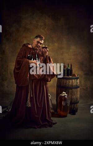 Mittelalterlicher Mönch in alter Kleidung hält Glas mit Cognac, Bier, Ale wieder Vintage Keller mit Holzfass und Flaschen oben. Abbey Production Stockfoto