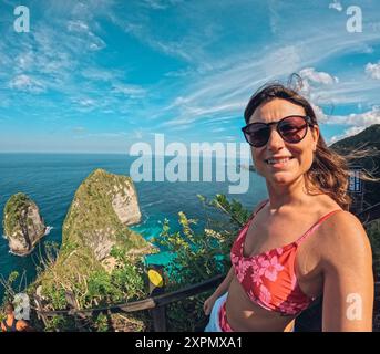 Eine Frau sitzt auf einem Felsvorsprung mit Blick auf den Ozean und trägt einen roten Bikini. Sie lächelt und blickt auf das Wasser Stockfoto