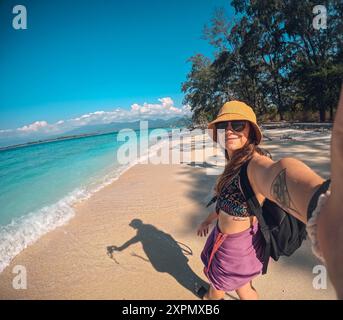 Eine Frau macht ein Selfie am Strand mit einem Mann hinter sich. Die Frau trägt einen gelben Hut und eine Sonnenbrille Stockfoto