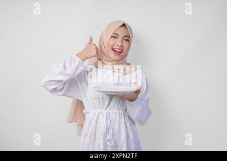 Aufgeregte junge asiatische muslimische Frau im Hijab, die leeren weißen Plattenkopierraum präsentiert, während sie die Daumen nach oben drückt Stockfoto