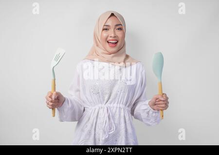 Porträt einer glücklichen asiatischen muslimischen Frau im Schleier-Hijab, die Spachtel und Küchenutensilien hält und fröhlich lächelt. Ramadan-Konzept Stockfoto