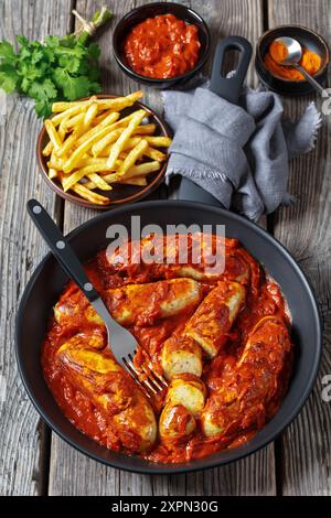 Deutsche Currywurst, Schweinebratwurst mit würziger und würziger Curry-Ketchup-Sauce in einer Pfanne auf grauem rustikalem Holztisch mit Pommes frites, Gabel, ver Stockfoto