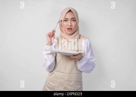 Verwirrte junge asiatische muslimische Frau in Hijab und Sahneschürze mit Löffel und leerem Teller mit einem Kopierraum darüber, und denkt hart, welche Speisenkarte zu essen ist Stockfoto