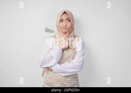 Nachdenkliche junge asiatische muslimische Frau in Hijab und Sahneschürze, die Spachtel und Küchenutensilien hält und sich intensiv Gedanken darüber macht, welche Speisenkarte man kochen soll Stockfoto