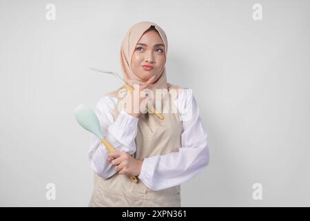 Nachdenkliche junge asiatische muslimische Frau in Hijab und Sahneschürze, die Spachtel und Küchenutensilien hält und sich intensiv Gedanken darüber macht, welche Speisenkarte man kochen soll Stockfoto