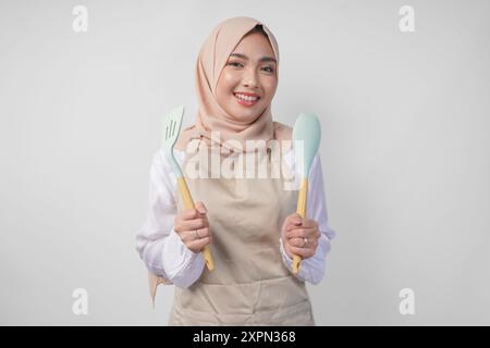 Aufgeregt junge asiatische muslimische Frau in einem Schleier Hijab und einer Creme-Schürze lächelt in die Kamera, während sie einen Spachtel und Küchenutensilien hält Stockfoto