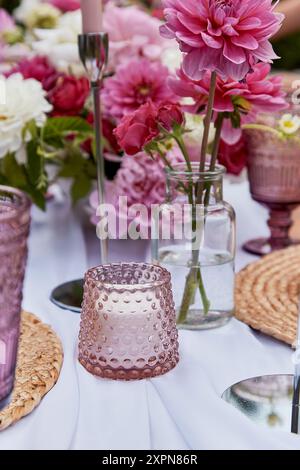 Elegante Kerze und rosafarbenes Glas-Votiv auf einem Tisch mit Blumenmotiven. Hochzeits- und romantische Abendthemen. Stockfoto