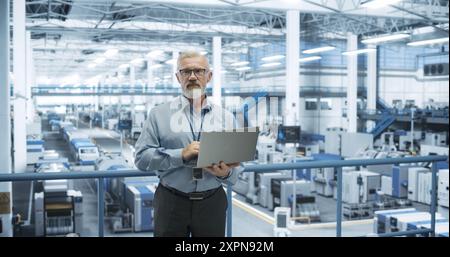 Porträt eines gutaussehenden Ingenieurs mittleren Alters mit Bart und Brille mit Laptop-Computer und Blick auf die Kamera. Männlicher Spezialist, der in einer modernen Elektronikfabrik arbeitet Stockfoto