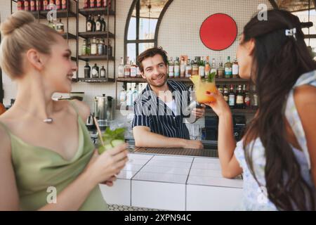 Zwei lächelnde junge Frauen treffen sich in der modernen Bar im Freien, genießen Cocktails und unterhalten sich fröhlich mit Mann, Barkeeper. Abendspaß Stockfoto