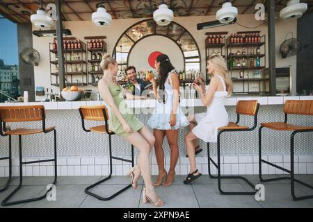 Drei lächelnde junge Frauen sitzen in der modernen Bar im Freien, genießen Cocktails und unterhalten sich fröhlich mit Mann, Barkeeper. Abendspaß Stockfoto