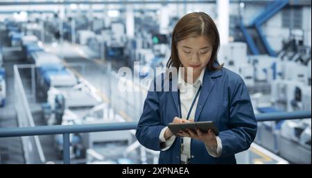 Junge asiatische Ingenieurin, die auf einer Plattform steht und einen Tablet-Computer in einer Elektronikfabrik benutzt. Maschinen werden gewartet, Spezialist überwacht den Fortschritt mithilfe der Online-Software Stockfoto