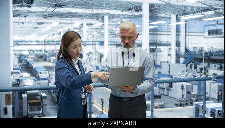 Männlicher Ingenieur und weiblicher Projektmanager, der auf einer Plattform steht, spricht, Laptop-Computer verwendet und die Produktion bei einer modernen automatisierten Elektronikfertigung mit KI-Robotern überwacht Stockfoto