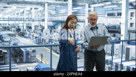 Ingenieur mittleren Alters und junger japanischer Techniker, die ein Gespräch führen und einen Laptop verwenden. Spezialisten für Überwachung, Analyse von Softwareberichten in einer modernen Elektronikfabrik Stockfoto
