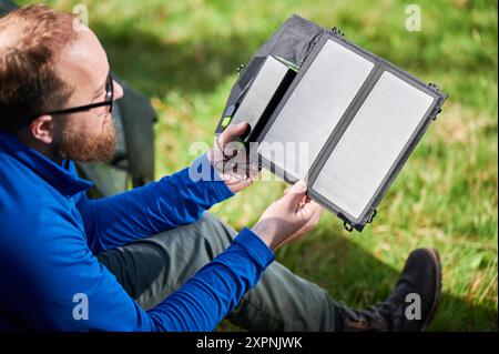 Der Mann sitzt neben dem Touristenzelt und hält ein tragbares Solarmodul-Ladegerät, um Sonnenlicht einzufangen und die Powerbank aufzuladen. Erneuerbare Energien bei Outdoor-Aktivitäten, die Technologie mit der natürlichen Umwelt verbinden. Stockfoto