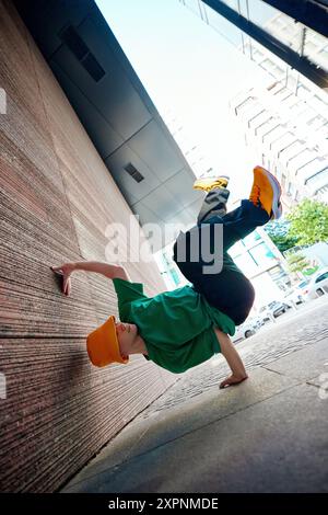 Breakdancer in grünem Hemd und orangefarbenem Eimerhut schlägt sich der Schwerkraft zu trotzen, balanciert mit einer Hand gegen die Stadtmauer, zeigt Agilität und Flair. Stockfoto