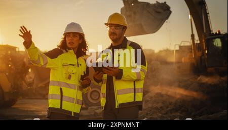 Filmische Golden-Hour-Aufnahme Der Baustelle: Kaukasischer Männlicher Bauingenieur Und Hispanische Inspektorin Sprechen Mit Tablet. Lkw, Bagger, Lader, Die Am Projekt Zur Landentwicklung Arbeiten. Stockfoto