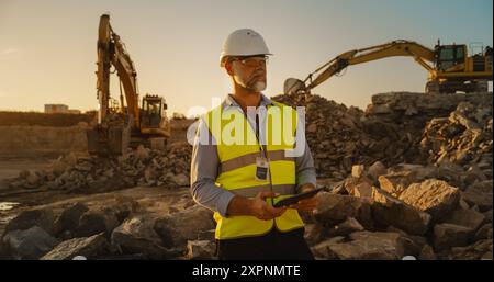 Kaukasischer Bauingenieur mit Schutzbrille und Tablet auf der Baustelle an Einem sonnigen Tag. Mann, Der Den Baufortschritt Überprüft. Bagger, Der Materialien In Das Flurförderzeug Lädt. Stockfoto