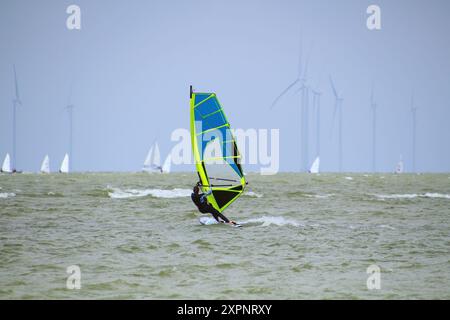 Junge Frau Windsurfen mit Segelbooten und Windturbinen am Horizont (Ijsselmeer, Niederlande) Stockfoto