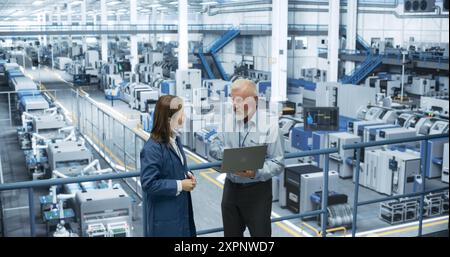Zwei multiethnische Ingenieure stehen auf einer Plattform, die Laptop-Computer in einer Elektronikfabrik verwenden. Maschinen werden gewartet, Spezialisten überwachen den Fortschritt mithilfe von Online-Software Stockfoto