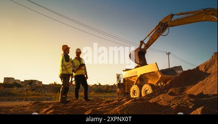 Kaukasischer Immobilieninvestor Mit Tablet Und Projektmanager Sprechen Auf Der Baustelle Des Wohngebäudes. Kollegen, Die Schutzhüte Tragen Und Heavy Machinery Bei Der Arbeit Zusehen. Sonniger Tag Stockfoto