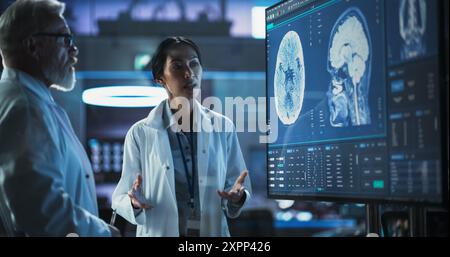 Neuroscience Laboratory Meeting: Asiatische Wissenschaftlerin und männliche kaukasische Chirurgin diskutieren MRT-Hirnscan auf TV-Display. Fachärzte Für Rehabilitationsmedikamente Nach Hirnschäden. Stockfoto