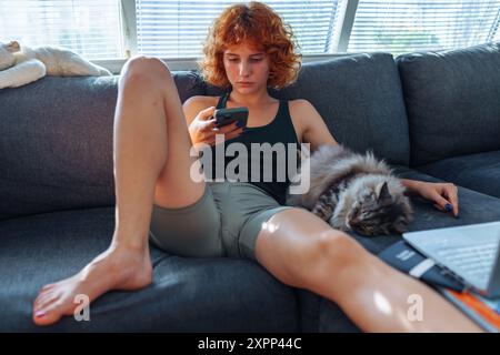 Porträt einer rothaarigen jungen Frau, die mit einem Laptop auf dem Sofa sitzt, eine graue Katze, die in der Nähe schläft Stockfoto