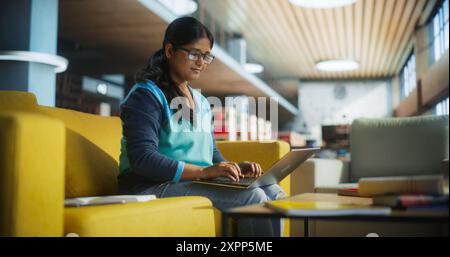 Smart Indian Female Studentin in Brille, die an Software Development Assignment in einer stillen öffentlichen Bibliothek arbeitet. Junge südasiatische Frau mit Laptop-Computer und Vorbereitung auf die Prüfung im College Stockfoto