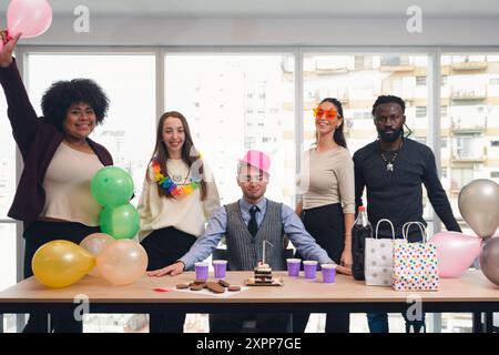 Kleine Versammlung von Bürokollegen, die einen der Kollegen zum Geburtstag feiern, Gruppe von multiethnischen Menschen mit Ballons, Geburtstagshüte und Stockfoto