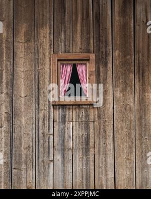 In einer alten Holzwand gibt es ein sehr kleines Fenster mit pinkfarbenen Vorhängen. Hohe Tauern. Österreich Stockfoto