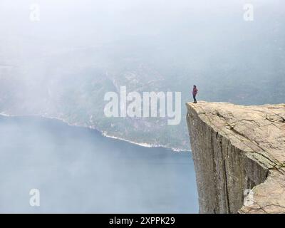 Eine einsame Figur steht triumphierend am zerklüfteten Rand von Preikestolen, umgeben von weichem Nebel und ruhigem Wasser. Stockfoto