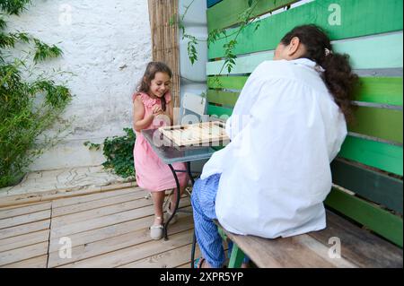 Ein fröhlicher Moment, in dem Mutter und Tochter gemeinsam Backgammon auf einer lebhaften und gemütlichen Terrasse spielen, wo Familienverbundenheit und Spaß gezeigt werden. Stockfoto