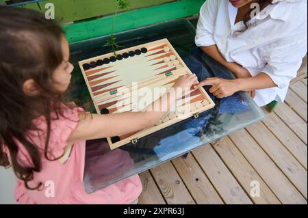 Mutter und Tochter spielen Backgammon auf einem Glastisch draußen. Ein Moment der Bindung, der Familienzeit und des strategischen Denkens. Stockfoto