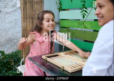 Glückliche Mutter und Tochter spielen zusammen Backgammon draußen im Garten. Familienbeziehungen, Spaß und Spielspaß. Stockfoto