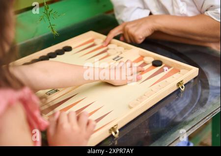 Nahaufnahme von zwei Leuten, die Backgammon auf einem Holzbrett spielen. Das Bild fängt den spannenden Moment eines Brettspiels in einer entspannten und lässigen Umgebung ein. Stockfoto