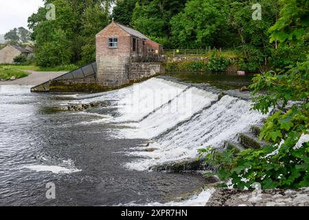 Grassington Yorkshire UK-27. Juli 2024. Eine ruhige Flussszene mit einem rustikalen Backsteingebäude und einem kaskadierenden Wehr inmitten von üppigem Grün. Stockfoto