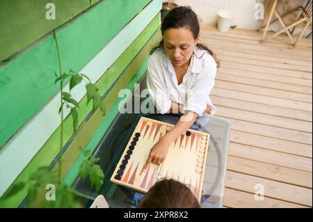 Eine Frau, die ein Spiel Backgammon auf ihrem Holzdeck genießt. Entspanntes und legeres Ambiente im Freien. Weibliche Spielerin, die einen Zug macht. Stockfoto