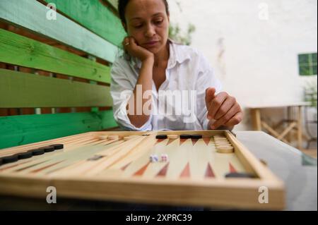 Eine Frau in einem weißen Hemd sitzt draußen und spielt Backgammon, fokussiert und nachdenklich, demonstriert strategisches Denken. Die Atmosphäre vermittelt ein entspanntes und Stockfoto