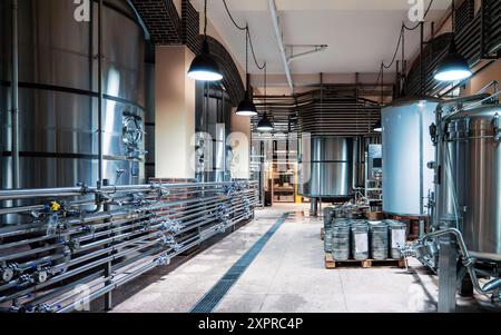 Geräumiges, modernes Brauereiinnere mit großen Edelstahltanks, komplizierten Rohrleitungen und stimmungsvoller Beleuchtung, die die Qualitätsstandards der Bierproduktion widerspiegelt. Stockfoto