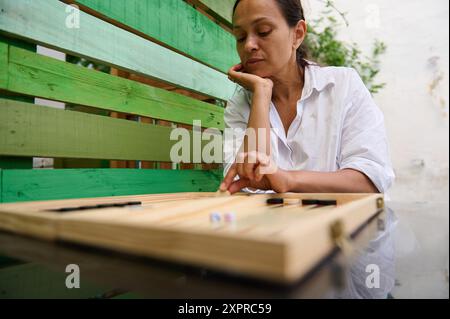 Eine Frau sitzt draußen in einem Garten und spielt Backgammon. Sie konzentriert sich auf das Brettspiel und genießt einen entspannenden Moment. Stockfoto